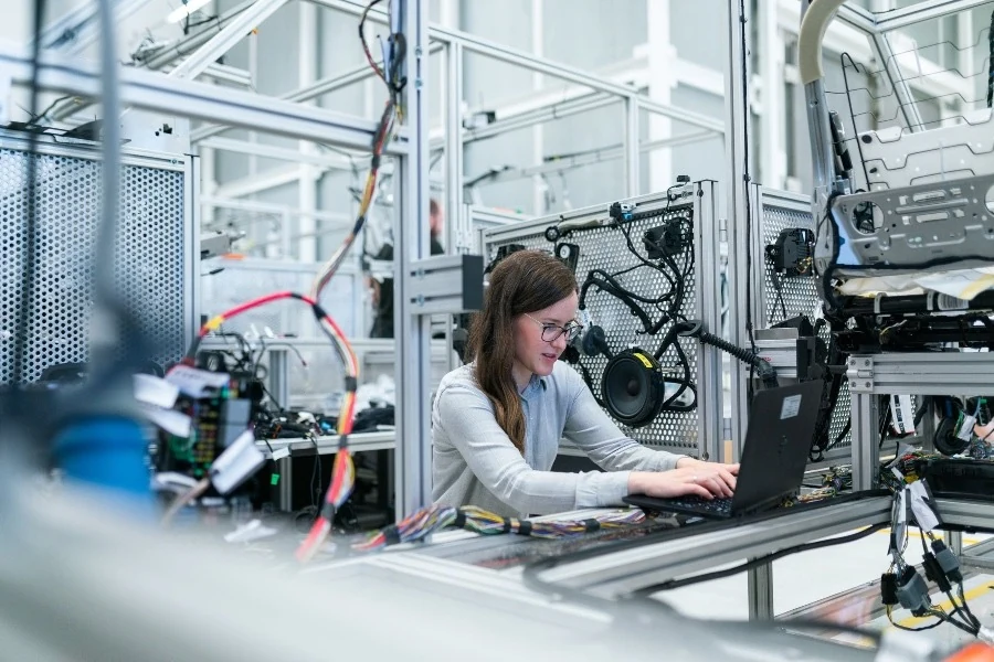 Una ingeniera trabaja en una laptop en un laboratorio de pruebas, rodeada de cableado y equipos electrónicos